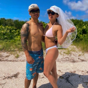 May include: A couple on a beach, the woman is wearing a white veil and a pink bikini top, the man is wearing blue swimming trunks and a white baseball cap.