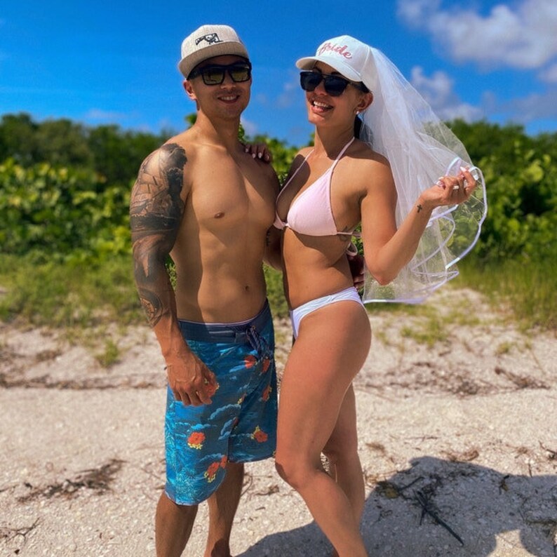 May include: A couple on a beach, the woman is wearing a white veil and a pink bikini top, the man is wearing blue swimming trunks and a white baseball cap.