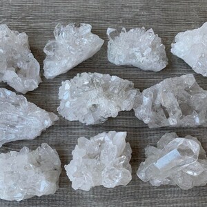 May include: A selection of clear quartz crystal clusters. The crystals have a natural, rough texture and vary in size and shape. They are arranged on a light-coloured wooden surface, showcasing their translucent quality.