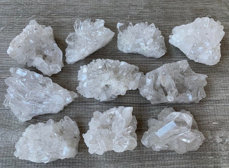 May include: A selection of clear quartz crystal clusters. The crystals have a natural, rough texture and vary in size and shape. They are arranged on a light-coloured wooden surface, showcasing their translucent quality.