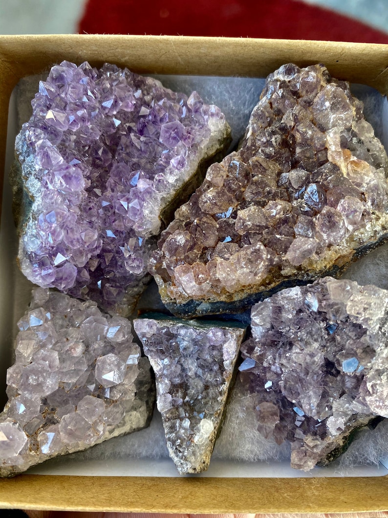 May include: A selection of amethyst crystal clusters displayed in a cardboard box. The crystals vary in shades of purple, from pale to rich hues, with visible crystalline formations. The box is lined with white padding, highlighting the natural beauty of the crystals.