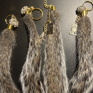Op de afbeelding: Vier bruine en grijze pluche sleutelhangers met gouden ringen en bedels. Elke sleutelhanger heeft een kleine, heldere strasssteen aan de bovenkant.