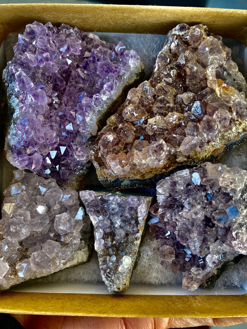 May include: A selection of amethyst crystal clusters displayed in a cardboard box. The crystals vary in shade from dark purple to pale lavender and are set on white padding. The crystals exhibit a sparkling, crystalline structure.