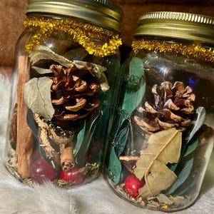 Könnte beinhalten: Zwei durchsichtige Einmachgläser mit dekorativen Gegenständen. Jedes Glas enthält einen Tannenzapfen, einen Zimtstange, rote Beeren und grüne Blätter. Die Gläser sind mit goldenen Deckeln und goldenem Lametta versehen.