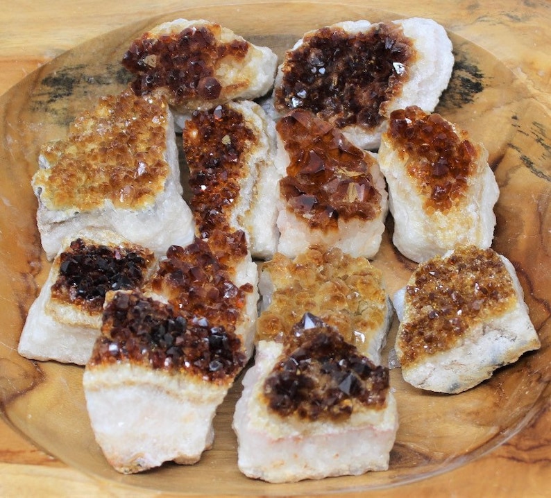 May include: A selection of raw citrine crystal clusters. The crystals exhibit amber and golden-brown tones, with a textured, sparkling surface. The citrine pieces are set against a white base, creating a striking contrast.