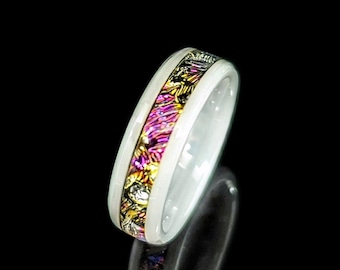 May include: White ceramic ring with a colorful, patterned inlay of pink, gold, and black. The ring's design features a central band of vibrant colors, framed by a smooth, white exterior. The ring is displayed against a black background.