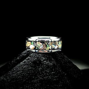 May include: A silver ring with a band of colorful, iridescent abalone shell fragments. The ring is set against a black background, highlighting the vibrant colors of the shell, including green, yellow, and blue.