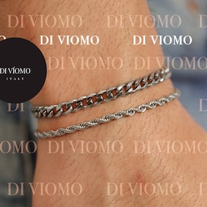 May include: A person's hand wearing three silver bracelets and two gold rings. The bracelets are a chain link, a rope chain, and a thinner chain. The rings are a simple band and a ring with a design on the top. The background is a blue denim fabric with the text "DI VIOMO" repeated.