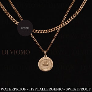 May include: A gold chain necklace with a pendant featuring the Libra zodiac symbol. The necklace is layered with a second, thinner gold chain. The text "WATERPROOF - HYPOALLERGENIC - SWEATPROOF" and "WWW.DIVIOMO.COM" are displayed below the image.