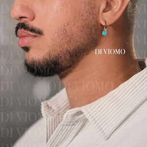 Puede incluir: Un hombre con una camisa blanca de cuello y un pendiente cuadrado turquesa. El pendiente está en el lado izquierdo de la imagen. El texto "DI VIOMO" es visible en la esquina inferior derecha de la imagen.