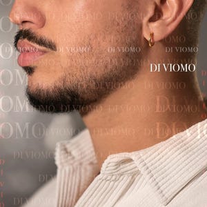 May include: A small, polished gold hoop earring worn in the earlobe. The earring features a simple, circular design with a smooth, reflective metallic finish. The text DI VIOMO is visible in the background and near the earring on the image.