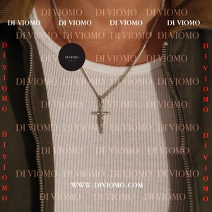 May include: A silver chain necklace with a silver cross pendant. The necklace is worn by a person wearing a white shirt and a green jacket. The text "DI VIOMO ITALY" is visible on a black circle tag hanging from the necklace.