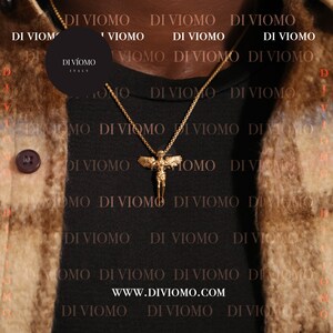 Puede incluir: Un collar de cadena dorada con un colgante que presenta una figura de &aacute;ngel alado. El collar lo lleva una persona que lleva una camisa negra con el texto "DI VIOMO" repetido en blanco.