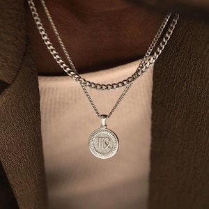 May include: A layered silver-toned necklace set featuring a delicate chain and a thicker curb chain. The pendant is a circular medallion with a textured border and a raised Virgo zodiac symbol in the center, crafted with a polished metallic finish.