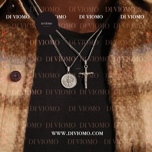 May include: Two silver chains with pendants. One pendant is a compass and the other is a cross. The chains are worn by a person with a brown leather jacket. The text "DI VIOMO" is repeated multiple times on the jacket and the text "WWW.DIVIOMO.COM" is visible in the center of the image.