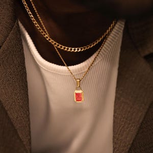 Pode incluir: Um colar de tom dourado com um pendente retangular que apresenta uma pedra preciosa vermelha vibrante no centro. O pendente pende de uma corrente dourada fina, sobreposta a uma corrente de elos dourada mais grossa sobre uma camisa branca.