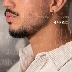 May include: A simple, polished silver-tone hoop earring worn in the earlobe. The earring features a thin, circular metal band design. The text DI VIOMO is visible in the background, and the earring is worn with a light-colored corduroy shirt.