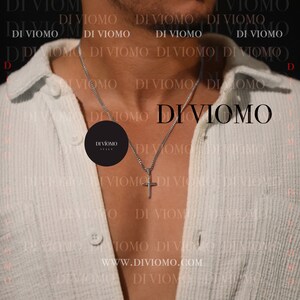 May include: A person wearing a white shirt with a silver chain necklace and a small cross pendant. The necklace has a black tag with the text "DIVIOMO ITALY" on it. The background is a white and black pattern with the text "DI VIOMO" repeated.