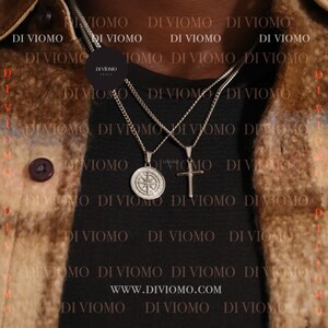 May include: Two silver chains with pendants, one a compass and the other a cross, hang from a person's neck. The chains are layered on top of each other. The background is a brown leather jacket with the text "DI VIOMO" repeated throughout. The text "WWW.DIVIOMO.COM" is also visible.