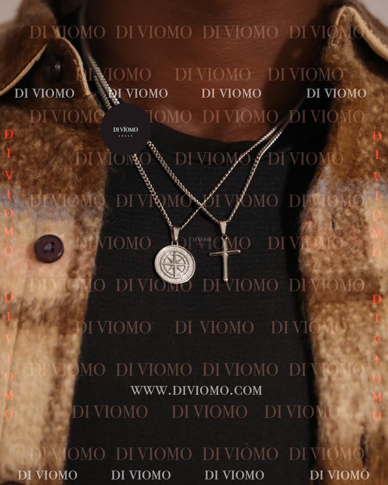 May include: Two silver chains with pendants, one a compass and the other a cross, hang from a person's neck. The chains are layered on top of each other. The background is a brown leather jacket with the text "DI VIOMO" repeated throughout. The text "WWW.DIVIOMO.COM" is also visible.