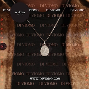 May include: A silver chain necklace with a silver pendant featuring a religious image. The necklace is worn by a person wearing a brown jacket. The background is a black and brown pattern with the text "DI VIOMO" repeated.