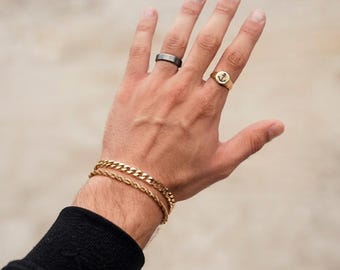 May include: A gold-toned curb chain bracelet, a gold-toned rope chain bracelet, a dark grey band ring, and a gold-toned signet ring featuring an anchor engraving. The text DI VIOMO ITALY is visible at the bottom of the image.