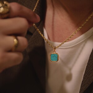 May include: A gold-toned rope chain necklace featuring a square pendant with a bright turquoise stone inlay. The pendant has a smooth, polished gold-colored metal frame that surrounds the square-cut turquoise center, which displays natural veining.