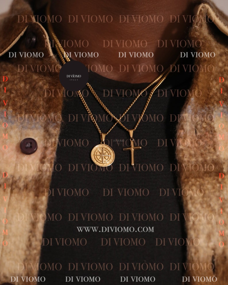 May include: Two gold chains with pendants, one a compass and the other a cross, worn on a person's neck. The chains are layered on top of each other. The background is a black shirt with the text "WWW.DIVIOMO.COM" and the words "DI VIOMO" repeated in a pattern.