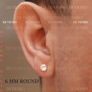 May include: A single round stud earring featuring a clear, faceted stone set in a gold-toned metal prong setting. The earring is positioned in an earlobe. The text 6 MM ROUND is visible in the lower left corner, along with the repeated watermark DI VIOMO.