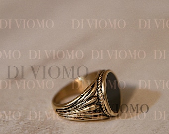 May include: A gold-toned metal signet ring featuring a smooth, circular black inlay at the center. The band displays a textured, fan-like pattern on the sides with a rope-style border surrounding the black stone. The text DI VIOMO is visible.