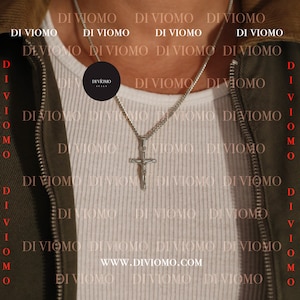 Puede incluir: Un collar de cadena de plata con un colgante de cruz de plata. El collar lo lleva una persona que lleva una camisa blanca y una chaqueta verde. El texto "DI VIOMO ITALY" es visible en una etiqueta circular negra que cuelga del collar.