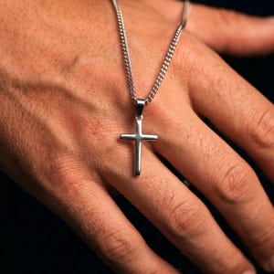 Puede incluir: Un colgante de cruz de metal plateado suspendido de una cadena de eslabones a juego. La cruz presenta un acabado liso y pulido con bordes rectos y limpios, y un diseño sencillo y sin adornos, que cuelga verticalmente de una pequeña argolla.