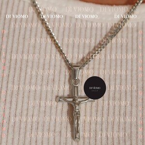 Puede incluir: Un collar de cadena de plata con un colgante de cruz de plata. La cruz presenta una figura de Jesucristo. El collar est&aacute; sobre un fondo blanco con el texto "DI VIOMO ITALY" repetido.