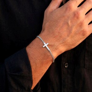 May include: A silver-toned metal chain bracelet featuring a small, centered cross charm. The delicate link chain wraps around the wrist, with the cross positioned horizontally as the focal point of the piece, creating a simple and clean aesthetic.
