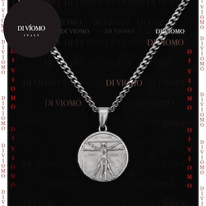May include: A silver-toned metal necklace featuring a circular pendant with a relief of the Vitruvian Man. The pendant is engraved with the text DI VIOMO and ITALY. The chain is a classic curb link style, and the piece is presented against a black background.