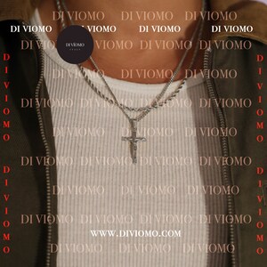 May include: A man wearing a white shirt with a silver chain necklace and a silver cross pendant. The necklace is layered with a second chain. The background is a green jacket with the text "DI VIOMO" repeated.