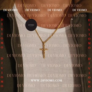 May include: A gold chain necklace with a gold cross pendant. The cross features a figure of Jesus Christ. The necklace is worn by a person wearing a white shirt and a green jacket. The text "www.diviomo.com" is visible on the shirt.