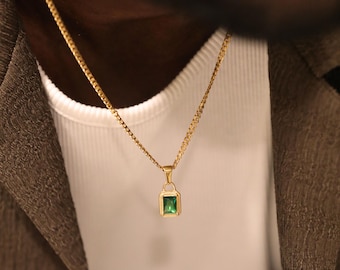 May include: A gold-toned chain necklace featuring a rectangular pendant with a vibrant emerald green gemstone set in a polished gold-colored frame. The pendant hangs from a delicate gold-toned link chain that rests against the chest area.