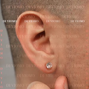 May include: A round, clear, faceted stud earring set in a metallic prong setting, positioned in the earlobe. The earring features a brilliant-cut stone that reflects light, creating a sparkling appearance against the skin of the ear.