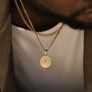 May include: A gold-toned layered necklace set featuring a circular pendant with an engraved compass rose design. The set includes a thicker curb chain and a thinner chain, both displaying a polished metallic finish and a classic link structure.