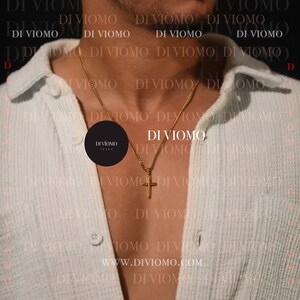 May include: A gold chain necklace with a small cross pendant. The necklace is worn by a person wearing a light blue shirt. The text "DI VIOMO" is repeated in the background.