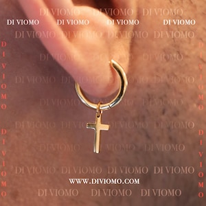 May include: Gold hoop earring with a small gold cross charm hanging from it. The earring is worn in a person's ear. The text "www.diviomo.com" is visible in the image.