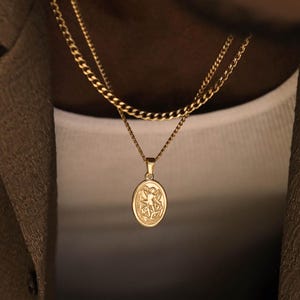 May include: A gold-toned layered necklace set featuring a thicker curb chain and a thinner chain with an oval pendant. The pendant displays a detailed embossed figure in a central relief design, suspended from a simple bail on a delicate link chain.