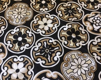 May include: Close-up of a black fabric with a repeating pattern of stylized floral medallions in white, tan, and black. The design has a vintage aesthetic. A ruler is visible along the bottom edge, with inch markings.