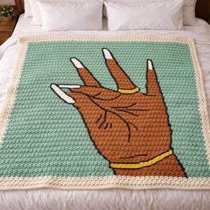 May include: A crocheted blanket featuring a brown hand with white-tipped nails, a gold ring, and a gold bracelet. The hand is set against a light green background with a cream-colored border. The blanket is on a white bed.