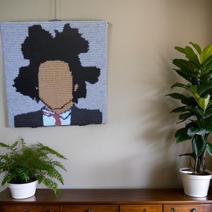 May include: A crocheted wall hanging featuring a stylized portrait with a black afro, tan face, and a dark suit with a red and blue tie. The artwork is set against a light blue background and is displayed on a wall.