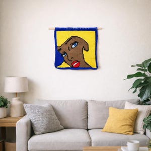May include: A colorful, handmade wall hanging featuring a stylized face. The artwork has a brown face with blue eyes and red lips, set against a yellow background with a blue border. It's mounted on a wooden dowel.