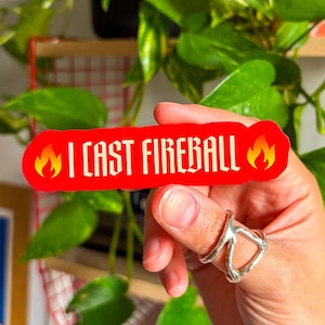 Peut inclure: Un autocollant rouge avec le texte "I CAST FIREBALL" en lettres blanches, avec un motif de flamme à chaque extrémité. L'autocollant est tenu dans une main avec une bague argentée, avec des plantes vertes en arrière-plan.