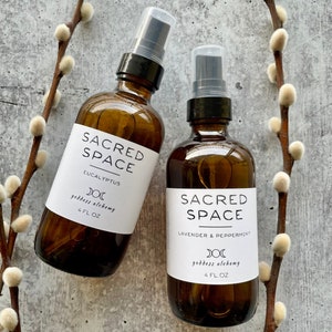 May include: Two amber glass bottles with spray tops, labeled "Sacred Space" with different scents: Eucalyptus and Lavender & Peppermint. Both bottles have a white label with a crescent moon symbol and the text "goddess alchemy" and "4 FL OZ".