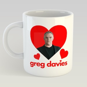 Puede incluir: Taza de cerámica blanca con un diseño de corazón rojo que presenta la cara de un hombre dentro del corazón. La taza también tiene dos corazones rojos más pequeños en el lateral y el texto "greg davies" en letras rojas en la parte inferior.